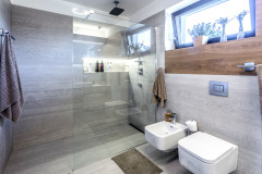 Sluncem prosvětlená koupelna kombinuje velkoformátový bezespárový obklad a dlažbu s dřevěným obkladem. Sprchový kout je oddělen minimalistickou skleněnou stěnou.