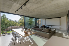 Společnému obývacímu prostoru dominuje atypické sezení, které je zčásti oboustranné.  Minimalistické vybavení a hladká cementová stěrka na podlaze jsou přizpůsobeny bydlení s malými dětmi