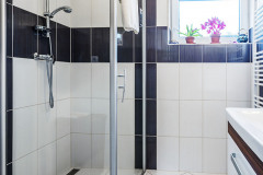 Prostorný sprchový kout a skříňka s umyvadlem v koupelně. Světlé dlaždice na zdech využívají kontrastu s tmavým odstínem
